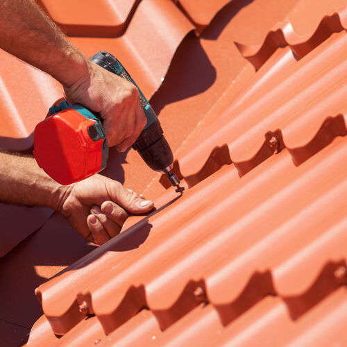 Worker on a roof with electric drill installing red metal tile on wooden house